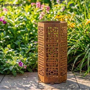 May include: A tall, hexagonal, rusted metal lantern with geometric cut-out designs. The lantern is placed outdoors among lush green plants and colorful flowers. The word "Lightinggo" is visible in the top left corner.