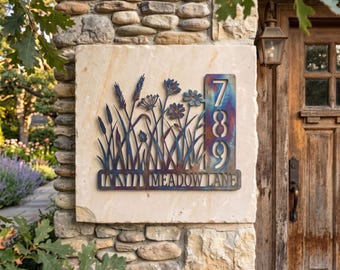 May include: A metal address sign featuring a wildflower meadow silhouette with the numbers 789 and the text MEADOW LANE. The sign has a heat-treated finish with blue, purple, and bronze tones, mounted on a light-colored stone wall surface.