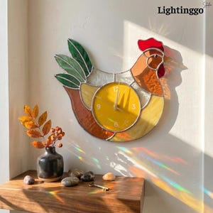 May include: A stained glass wall clock in the shape of a chicken. The clock face is yellow with black numbers and hands. The chicken has a red comb, orange and brown body, and green tail feathers. The clock is mounted on a white wall. The text "Lightinggo" is in the upper right corner.
