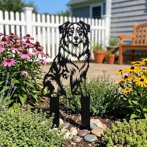 Peut inclure: Un piquet de jardin en métal noir représentant la silhouette détaillée d'un chien assis. Le chien a un pelage touffu et est placé dans un jardin avec des fleurs et une clôture blanche. Le piquet est conçu pour être planté dans le sol.