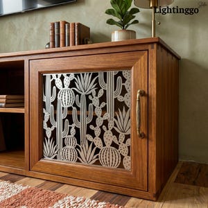 May include: A wooden media console featuring a cabinet door with a laser-cut metal panel displaying a detailed cactus and succulent pattern. The cabinet includes a vertical brass-toned handle and an open shelf section containing several books.