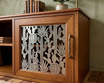 Puede incluir: Un mueble multimedia de madera con una puerta de armario que presenta un panel de metal cortado con láser con un patrón detallado de cactus y suculentas. El armario incluye un tirador vertical de tono latón y un estante abierto con libros.