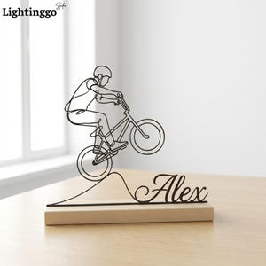 Könnte beinhalten: Schwarze Metallskulptur eines Radfahrers auf einem BMX-Rad, der einen Wheelie macht. Die Skulptur ist auf einem hellen Holzsockel montiert, mit dem Namen "Alex" in Schreibschrift. Das Lightinggo-Logo befindet sich oben links.