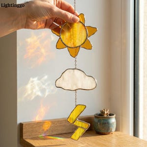 May include: A stained glass sun, cloud, and lightning bolt hanging decoration. The sun is yellow with a starburst design, the cloud is white, and the lightning bolt is yellow. The decoration is suspended by a chain.