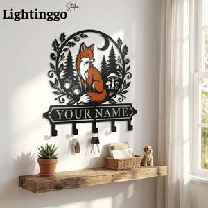May include: A black metal key holder with a fox design, forest elements, and the text "YOUR NAME." The key holder has four hooks and is mounted on a white wall. A wooden shelf below holds keys, a plant, and a small dog figurine.
