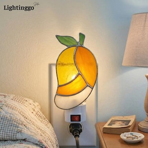 May include: A stained glass night light shaped like a mango, featuring yellow, orange, green, and white glass. The night light is plugged into a wall outlet and has a red on/off switch. The brand name "Lightinggo" is visible in the top left corner.