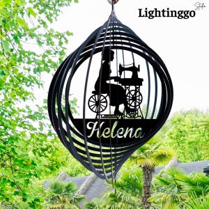 May include: Black metal wind spinner featuring a silhouette of a person sewing on a vintage sewing machine. The name "Helena" is displayed below the silhouette. The wind spinner has the text "Lightinggo" at the top.