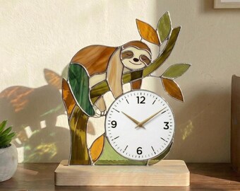 May include: A decorative stained glass clock depicting a sloth on a tree branch. The clock face is white with black numerals and gold hands. The sloth and tree are in brown, green, and orange, set on a wooden base.