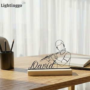 May include: A black line art sculpture of a person playing a flute, with the name "David" in cursive script on a wooden base. The sculpture is on a wooden table, with pencils in a black holder to the left.