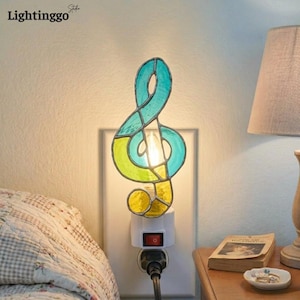 May include: A stained glass night light shaped like a treble clef, featuring teal, lime green, and yellow glass segments held by metal solder lines. The light is plugged into a white wall outlet and emits a warm glow from a small bulb.