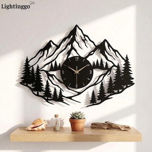 May include: A black metal wall clock featuring a laser-cut silhouette of a mountain range and pine forest. The circular clock face is integrated into the center of the design, displaying gold-colored hands and numerals at the 12, 3, 6, and 9 positions.