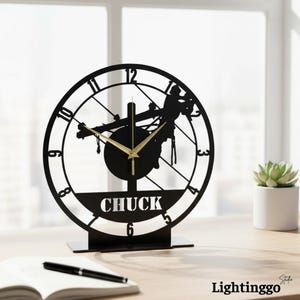 May include: Black metal desk clock with a lineman silhouette design. The clock features gold-colored hands and the name "CHUCK" at the bottom. The clock is on a black base and sits on a wooden surface.