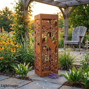 Puede incluir: Una farola de jardín rectangular de metal color óxido con diseños recortados de flores y gatos. La farola está rodeada de flores y vegetación, con una pérgola y una silla de madera al fondo. El texto "Lightinggo" está en la parte inferior.