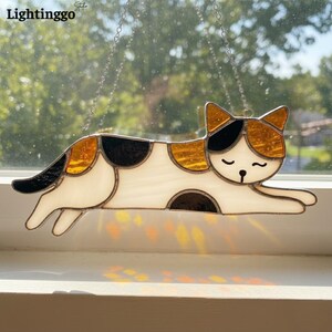 May include: Stained glass cat suncatcher with a white body, black and gold spots, and a black tail. The cat is in a relaxed pose, with its eyes closed. The suncatcher is hanging from a chain. The word "Lightinggo" is in the top left corner.
