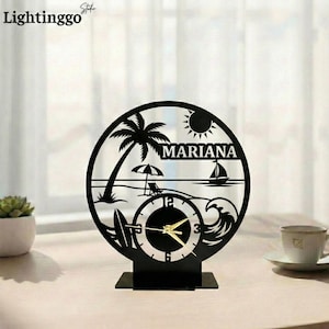 May include: Black laser-cut wooden clock with a beach scene design. The clock features a palm tree, sun, sailboat, and the name "MARIANA". The clock hands are gold, and the clock sits on a rectangular base.