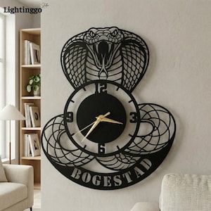 May include: Black metal wall clock featuring a cobra design. The clock face has gold hands and the numbers 12, 3, 6, and 9. The word "BOGESTAD" is at the bottom of the clock. The clock is mounted on a light-colored wall.