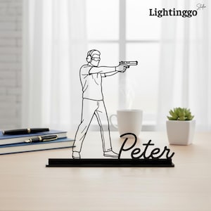 Puede incluir: Silueta negra cortada con láser de una persona apuntando con una pistola, con gafas protectoras y auriculares. La figura está montada sobre una base negra con el nombre "Peter" en cursiva. El fondo incluye una taza de café, una suculenta y el texto "Lightinggo".