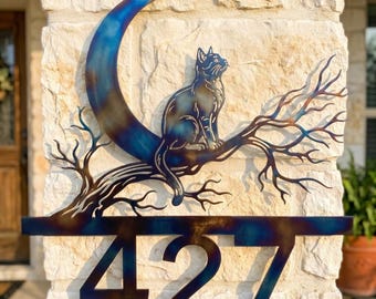 May include: Metal house number sign with a cat on a branch and a crescent moon. The sign shows the number 427 in a stylized font. The sign has a blue and bronze colour scheme and is mounted on a stone wall. The sign is a decorative item.
