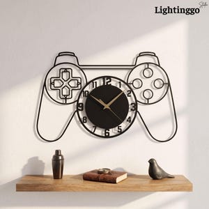 May include: A black metal wall clock shaped like a video game controller. The clock features a circular centre with black numbers from 1 to 12 and thin gold-coloured hour and minute hands. The frame outlines the buttons and joysticks of a controller.