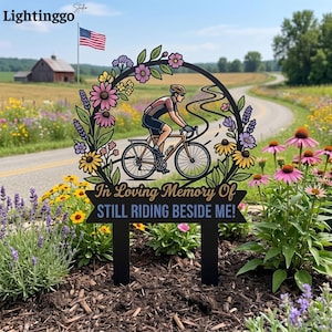Puede incluir: Estaca de jardín de metal negro con un ciclista en una carretera sinuosa, rodeado de una corona floral. El texto dice "In Loving Memory Of STILL RIDING BESIDE ME!". Una bandera americana ondea al fondo.