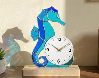 May include: A decorative seahorse clock with a white clock face and gold hands. The seahorse is made of stained glass in shades of blue and turquoise, mounted on a wooden base. The clock is a unique home decor item.