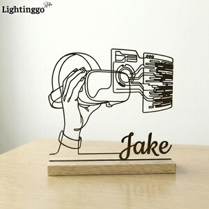 May include: A black metal VR headset design with the name "Jake" in cursive script, mounted on a wooden base. The image is a line drawing of a hand holding the headset. The logo "Lightinggo" is in the top left corner.