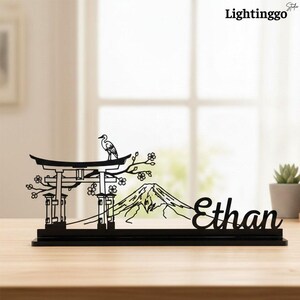 Könnte beinhalten: Schwarze, lasergeschnittene Holz-Schreibtischdekoration mit einem japanischen Torii-Tor, dem Berg Fuji und einem Kranich auf einem Ast. Der Name "Ethan" ist ebenfalls enthalten. Die Dekoration steht auf einer Holzoberfläche.