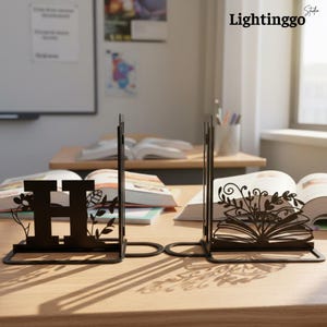 May include: Black metal bookends with decorative floral and open book designs. One bookend features the letter "H" with floral accents. The other bookend has an open book design with leaves and vines. The background shows open books on a wooden desk.