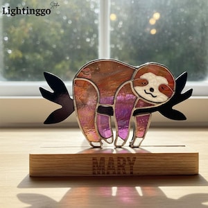 Può includere: Una statuetta di bradipo in vetro colorato nei toni dell'arancione, rosa e viola, appollaiata su un ramo nero. Il bradipo è montato su una base di legno con il nome "MARY" inciso. Il logo "Lightinggo" si trova nell'angolo in alto a sinistra.