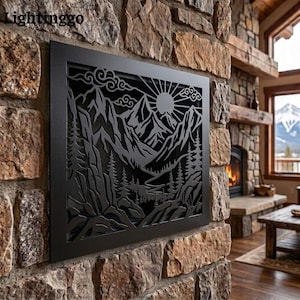 May include: A rectangular black metal wall art panel featuring a laser-cut design of a mountain range, a sun with radiating rays, stylized clouds, and a forest of pine trees. The piece is mounted on a textured stone wall surface.