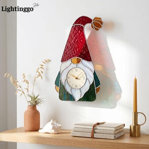 Könnte beinhalten: Eine Wanduhr aus Buntglas in Form eines Gnoms mit hohem rotem Hut, weißem Bart und grünem Sockel. Das Zifferblatt ist kreisförmig mit goldenen Zeigern und schwarzen Ziffern. Das Glas besteht aus texturierten roten, weißen und grünen Elementen.