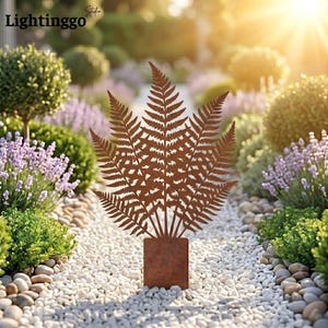 May include: A decorative metal fern sculpture with a brown, weathered finish. The fern is mounted on a square base and stands in a garden path lined with white stones, surrounded by greenery and flowers.
