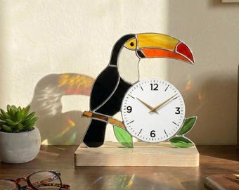 May include: A decorative toucan clock with a black body, a yellow, orange, and red beak, and a white clock face. The clock is perched on a wooden base and features gold-coloured clock hands. The clock is a unique home decor item.