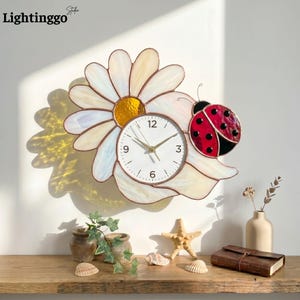May include: A daisy-shaped wall clock with a white face and gold hands. The clock is surrounded by a stained glass flower with white and yellow petals. A red and black ladybug is perched on the flower. The clock is labeled "Lightinggo Studio".