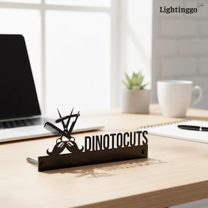 Könnte beinhalten: Schwarzes Metall-Friseurschild mit dem Text "DINOTOCUTS". Das Schild zeigt einen Kamm, eine Schere und ein Schnurrbart-Design. Das Schild steht auf einem Holztisch mit einem Laptop, einer Kaffeetasse und einem Notizblock im Hintergrund.