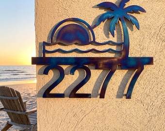 Puede incluir: Un letrero de pared de metal con los números 2277 en fuente negrita, coronado con la silueta de una palmera, un sol poniente y olas del mar. El metal tiene un acabado tratado térmicamente con degradados de color azul, morado y bronce.