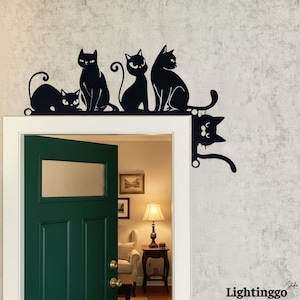 May include: Black metal cat silhouette decor, featuring five cats in various poses, perched above a green door. The cats are arranged in a row, with one cat hanging off the side of the door frame. The decor adds a whimsical touch to any doorway.