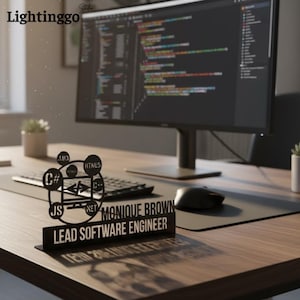 May include: A black desk nameplate with the text "MONIQUE BROWN LEAD SOFTWARE ENGINEER". The nameplate features a cut-out design with programming language symbols like C#, HTML5, and Java. A computer monitor and keyboard are in the background.