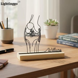 Könnte beinhalten: Eine schwarze Metall-Kniegelenk-Skulptur auf einem Holzsockel mit dem Namen "Williams" in Schreibschrift. Die Skulptur ist eine Strichzeichnung des Kniegelenks. Der Hintergrund umfasst eine kleine Topfpflanze und Bücher.