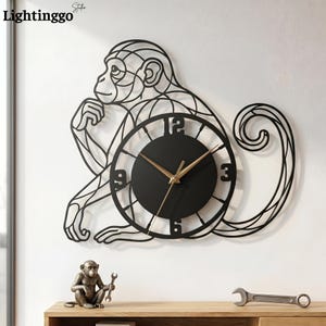 Puede incluir: Un reloj de pared de metal negro con forma de mono sentado, con un centro circular que presenta manecillas doradas y los números 3, 6, 9 y 12. El diseño del mono está compuesto por líneas finas interconectadas que forman una silueta geométrica.