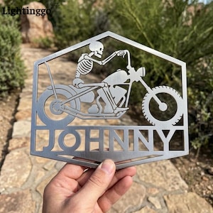 May include: A metal sign depicting a skeleton on a motorbike. The sign is hexagonal and has the name "JOHNNY" cut out at the bottom. The sign is held by a hand.