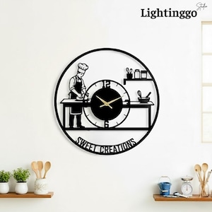 May include: Black metal wall clock featuring a chef preparing food, with the text "Sweet Creations". The clock has a circular design with a clock face and gold-colored hands. The brand name "Lightinggo" is at the top.
