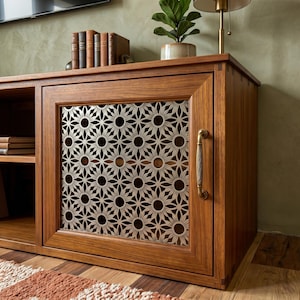 Op de afbeelding: Een houten mediaconsole met een kastdeur voorzien van een metalen, lasergesneden bloemmotief en een verticale messing handgreep. De console heeft een open schapgedeelte aan de linkerkant en een gladde afwerking met houtnerf.