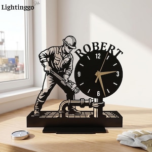 Può includere: Un orologio da tavolo nero con la sagoma di un operaio con casco che usa una chiave inglese su un tubo. Il quadrante circolare mostra il nome ROBERT in alto, con lancette dorate per ore, minuti e secondi su uno sfondo nero.