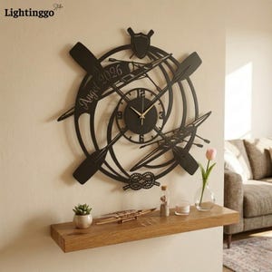 May include: Black metal wall clock with a nautical theme. The clock features crossed oars, a boat, and a decorative knot. The clock face has gold hands and the text "Angel 2026".