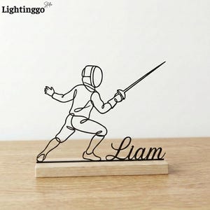 May include: A black metal sculpture of a fencer in action, mounted on a rectangular wooden base. The sculpture is a continuous line drawing, with the name "Liam" written in cursive script below the fencer.
