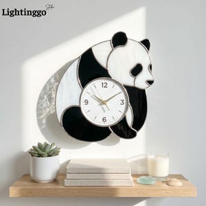 May include: A decorative panda-shaped wall clock. The clock face is white with gold hands, set within the panda's black and white body. The clock is mounted on a white wall. The brand name "Lightinggo" is in the top left corner.