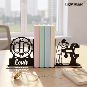 Op de afbeelding: Een paar boekensteunen van zwart metaal met wetenschappelijke thema's. De ene boekensteun toont een cirkelvormig deeltjesversnellerontwerp met de naam Louis in cursief schrift, de andere een silhouet van een wetenschapper bij een microscoop.