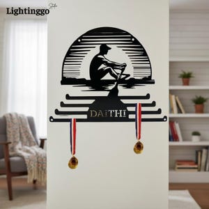 Puede incluir: Arte de pared de metal negro que representa la silueta de un remero en un bote bajo un sol estilizado. La pieza de arte incluye un soporte para medallas con el nombre "DAITHI" y dos medallas con cintas rojas, blancas y azules.