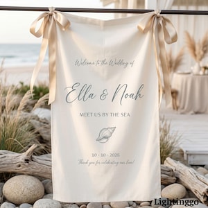 Könnte beinhalten: Ein cremefarbenes Hochzeits-Willkommensschild mit goldenen Schleifen. Auf dem Schild steht "Willkommen zur Hochzeit von Ella & Noah, Treffen Sie uns am Meer" mit einer Muschelgrafik und dem Datum 10.10.2026. Unten steht "Vielen Dank, dass Sie unsere Liebe feiern!"